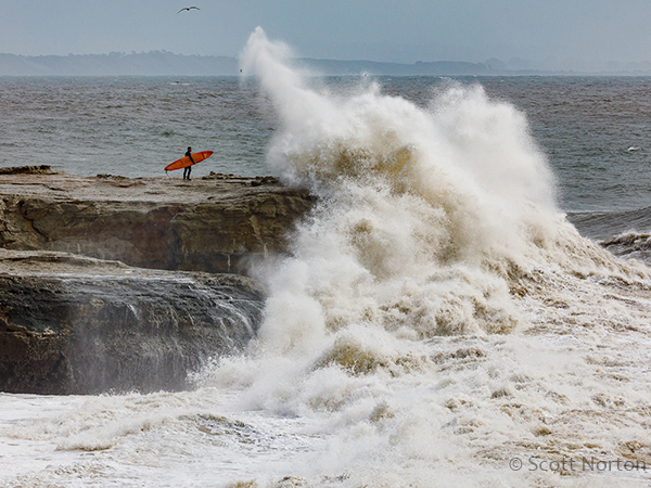 surfer standing on rock in santa cruz as large wave crashes by scott norton